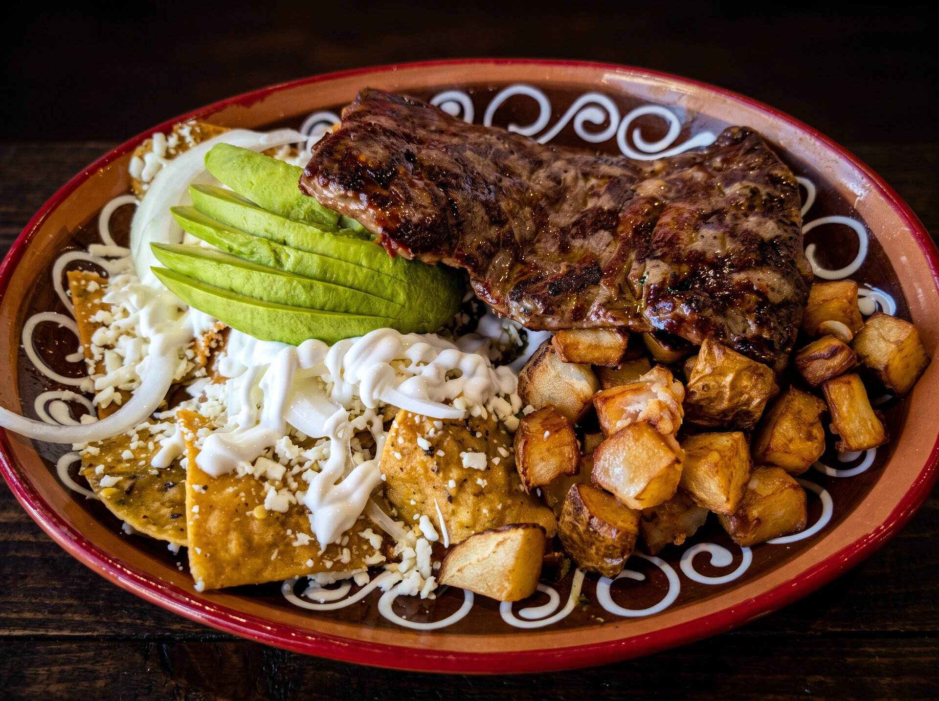 Chilaquiles topped with grilled carne asada, avocado, crema and potatoes on a red clay plate