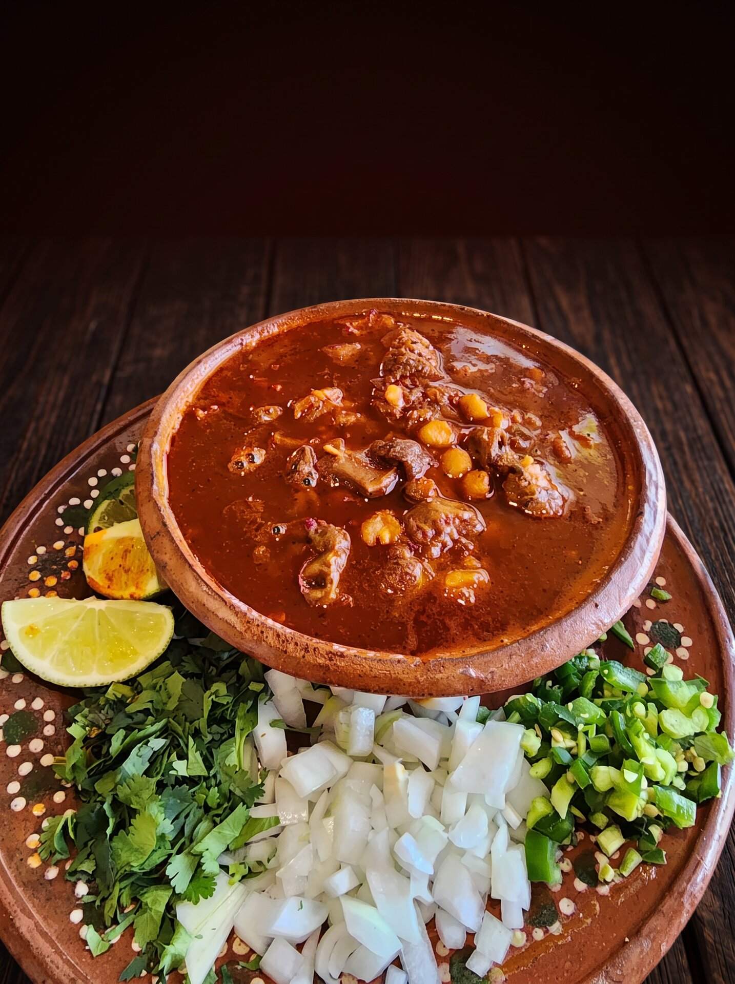 A clay bowl of red pozole with hominy, beside cilantro, onion and lime