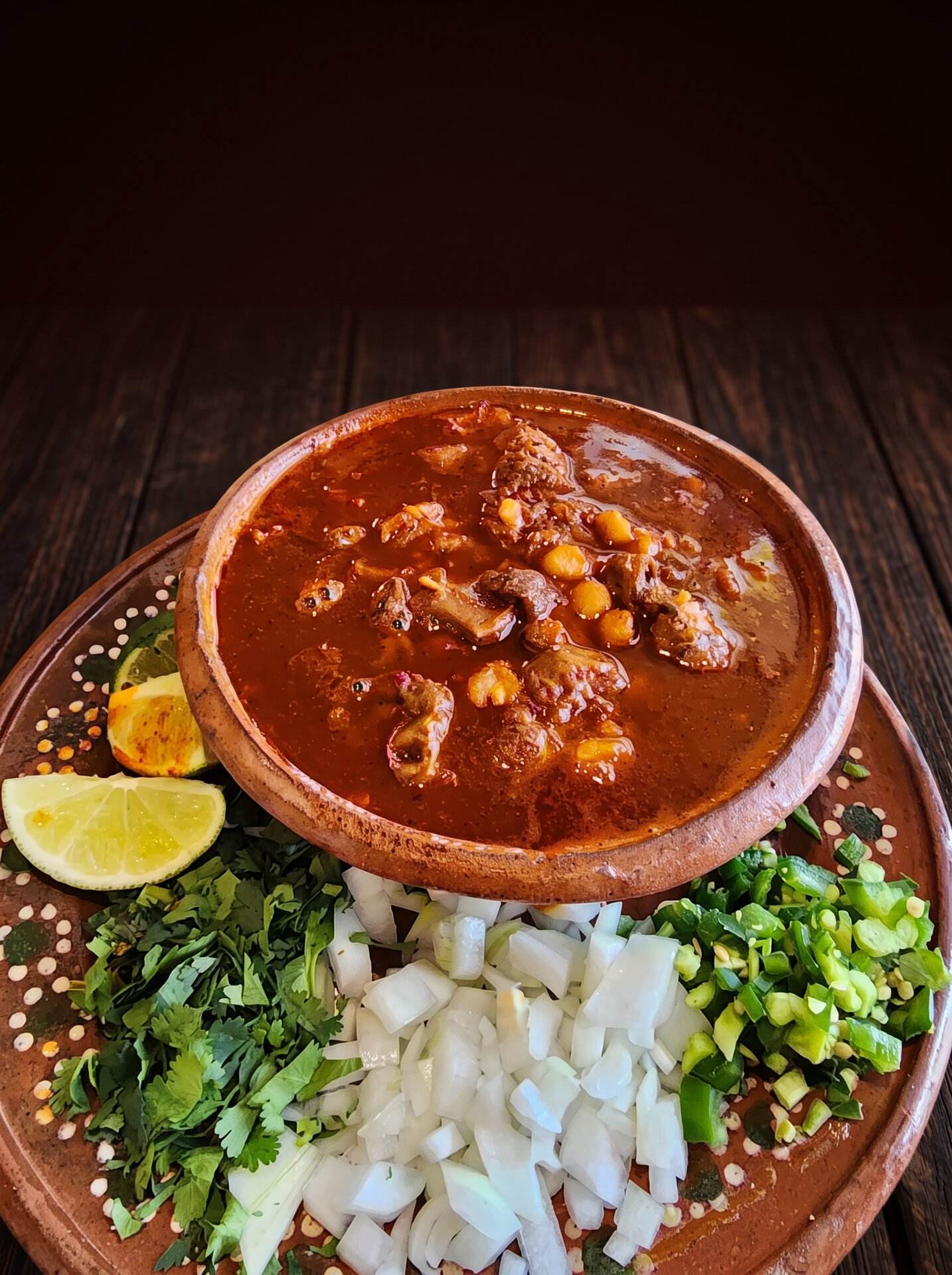 A clay bowl of red pozole with hominy, beside cilantro, onion and lime