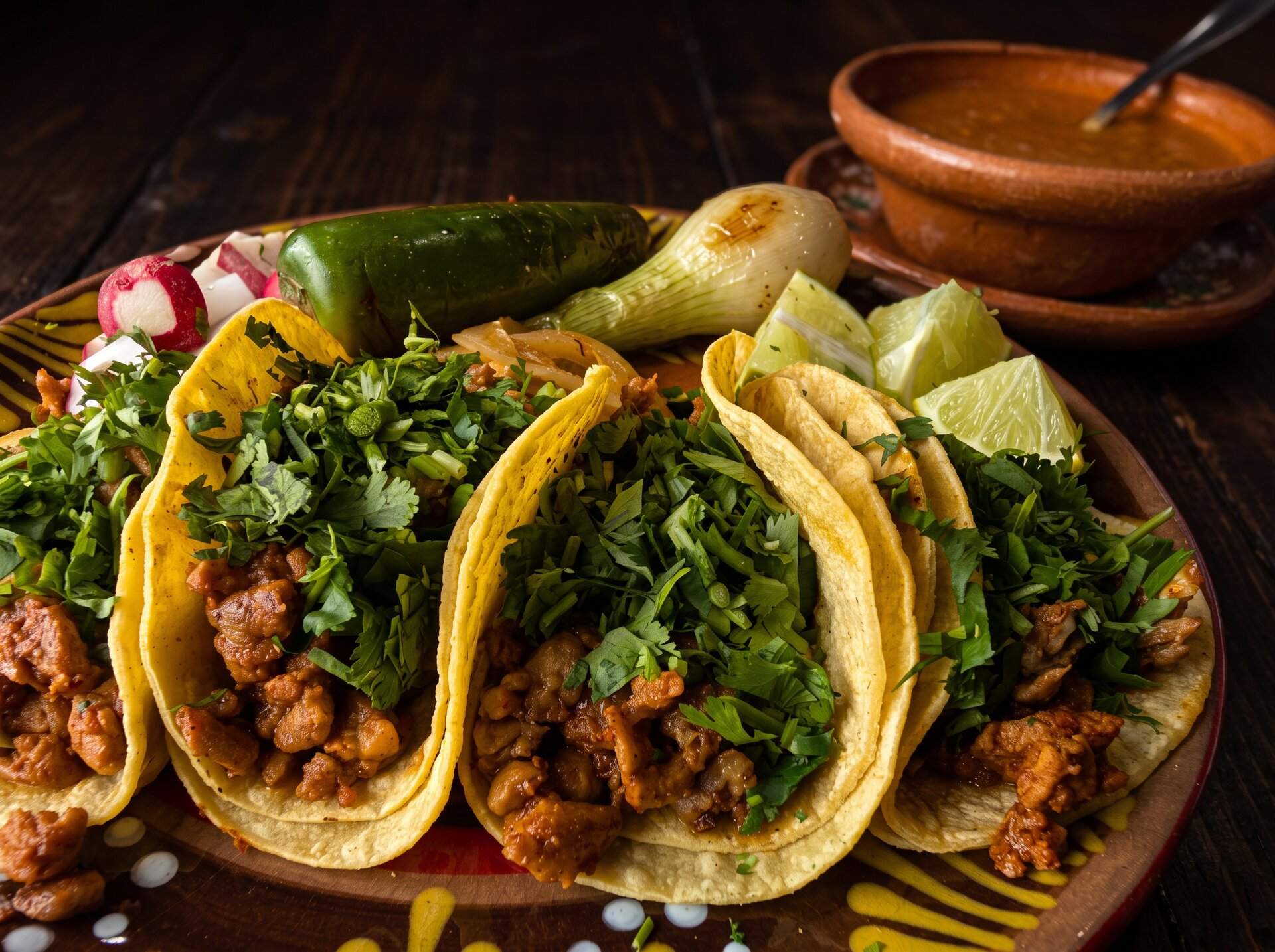 Three tacos with cilantro and onion on a clay plate with a grilled chile toreado and radish