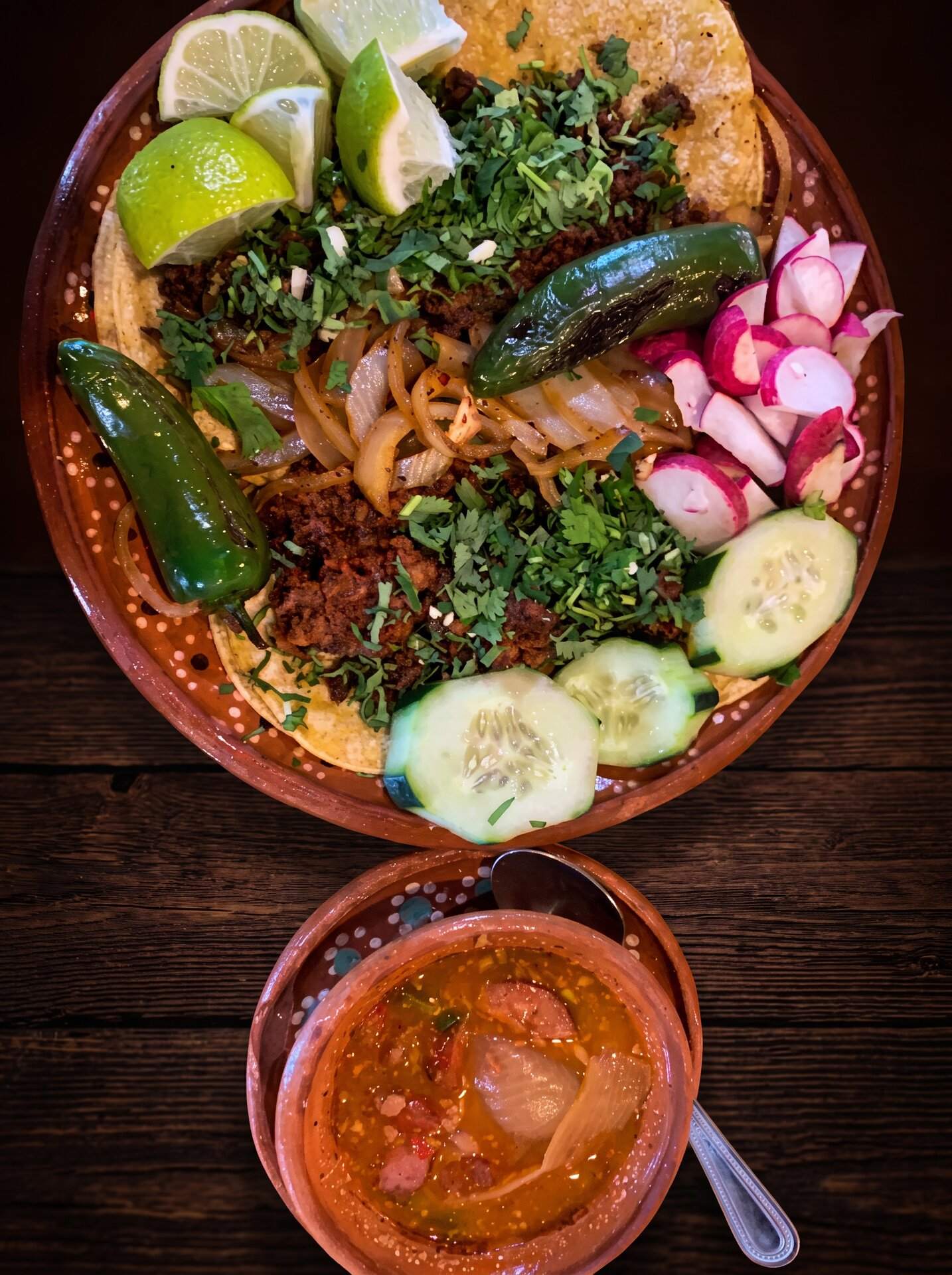 Four tacos with cilantro, radish and cucumber over a clay plate, salsa alongside